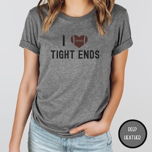 May include: A heather gray t-shirt with the text "I ❤️ TIGHT ENDS". The heart symbol is replaced with a brown football. The shirt has short sleeves and is being worn by a person.