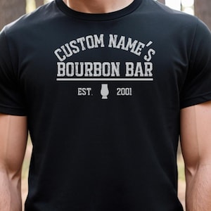 May include: Black t-shirt with white text that reads "CUSTOM NAME'S BOURBON BAR" above a line, with "EST. 2001" below, and a bourbon glass graphic. The shirt is a short-sleeved crew neck.