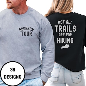 Op de afbeelding: Twee sweatshirts, één grijze en één zwarte. Het grijze sweatshirt heeft "BOURBON TOUR" op de voorkant gedrukt. Het zwarte sweatshirt heeft "NOT ALL TRAILS ARE FOR HIKING" en een Kentucky-omtrek. Een afbeelding toont "38 DESIGNS".