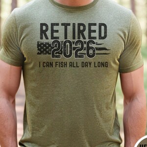 May include: Olive green t-shirt with the text "RETIRED 2026" above a stylized American flag and the words "I CAN FISH ALL DAY LONG". The shirt is a short-sleeved crew neck.