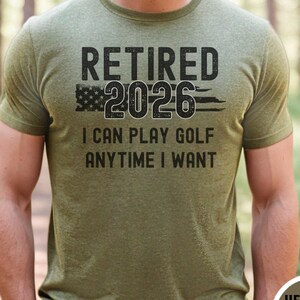 May include: Olive green t-shirt with the text "RETIRED 2026" above "I CAN PLAY GOLF ANYTIME I WANT". The number 2026 is stylized with an American flag design. The shirt is a short-sleeved crew neck.