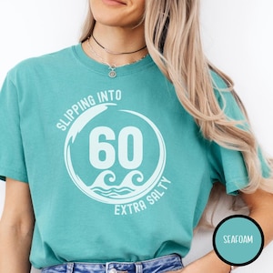 May include: Seafoam green t-shirt with the text "Slipping Into 60 Extra Salty" in white. The design includes a circle with the number 60 and wave graphics. The shirt is a solid color and has a round neckline.