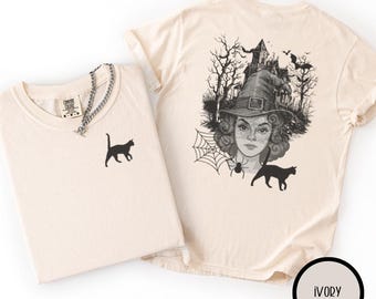 May include: Ivory-colored t-shirts. One shirt features a black cat silhouette, the other a witch illustration with a spiderweb, bats, and a haunted house. The shirts are folded, with a silver chain necklace visible.
