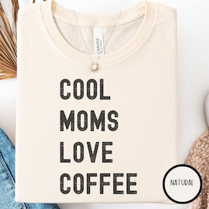 Puede incluir: Camiseta color crema con el texto "COOL MOMS LOVE COFFEE" en negrita negra. La camiseta tiene cuello redondo y mangas cortas. Una pequeña etiqueta circular "NATURAL" es visible en la parte inferior derecha.