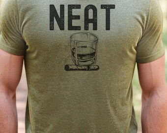 May include: Olive green t-shirt with the word "NEAT" in bold, black letters. Below the text is a graphic of a rocks glass with whiskey and a cigar. The shirt is short-sleeved.