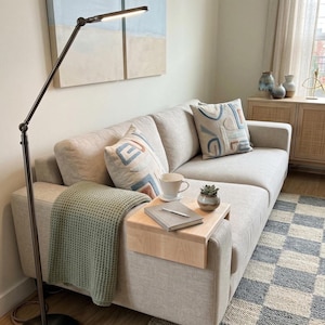 May include: A contemporary living room with a light beige sofa, patterned cushions, and a sage green throw. A wooden side table holds a mug, book, and plant. A floor lamp and abstract artwork create a comfortable atmosphere.