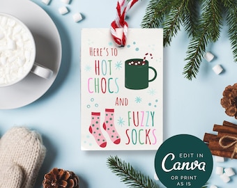 Pode incluir: Um cartão branco com um design festivo que apresenta uma caneca de chocolate quente, um par de meias felpudas e o texto "Here's to *HOT* CHOCS and *FUZZY SOCKS". O cartão está pendurado em um cordão vermelho e branco. O fundo é uma superfície azul claro com marshmallows, paus de canela, pinhas e um ramo de pinheiro.