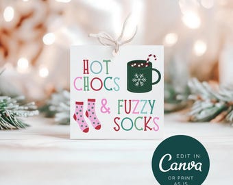 Pode incluir: Uma etiqueta quadrada branca com o texto "HOT CHOCS & FUZZY SOCKS" em rosa, verde e vermelho. A etiqueta apresenta ilustrações de uma caneca de chocolate quente, um floco de neve e meias rosa. Um círculo verde escuro com o texto "EDIT IN Canva OR PRINT AS IS" está no canto inferior direito.