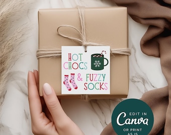 Pode incluir: Uma caixa de presente embrulhada com uma etiqueta que diz "Hot Chocs & Fuzzy Socks". A etiqueta apresenta uma caneca de chocolate quente, flocos de neve e meias rosa com bolinhas brancas. O presente é amarrado com barbante.