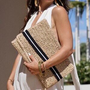 May include: A woven straw clutch with a black and white stripe detail. The clutch is rectangular and appears to be handmade. The bag is held in front of a person wearing a white jumpsuit and sunglasses. Gold bracelets and earrings are also visible.