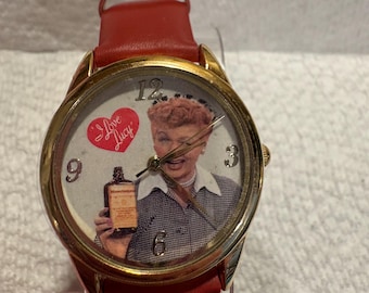 May include: An analog wristwatch with a gold-tone metal case and a red leather strap. The watch face features a portrait of Lucille Ball, a red heart with the text I Love Lucy, and gold-tone numerals at the 3, 6, 9, and 12 o'clock positions.