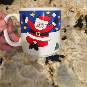 Puede incluir: Una taza de cerámica blanca con un gráfico de Santa Claus con traje rojo y cinturón negro, saltando sobre un fondo azul con estrellas amarillas. El diseño incluye un suelo blanco nevado y un pequeño pino verde en la parte inferior derecha.