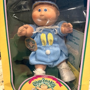 May include: A 1985 Cabbage Patch Kids Preemie doll in original packaging. The doll has a round face, blue eyes, and wears a light blue outfit with yellow gingham bunny ears and a white pom-pom tail. It includes a birth certificate and adoption papers.