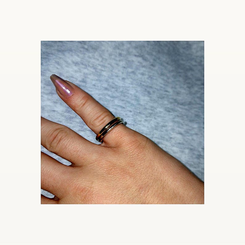 May include: A silver ring with a double band design is worn on a finger. The ring has a polished finish, reflecting light. The background is a soft grey fabric.