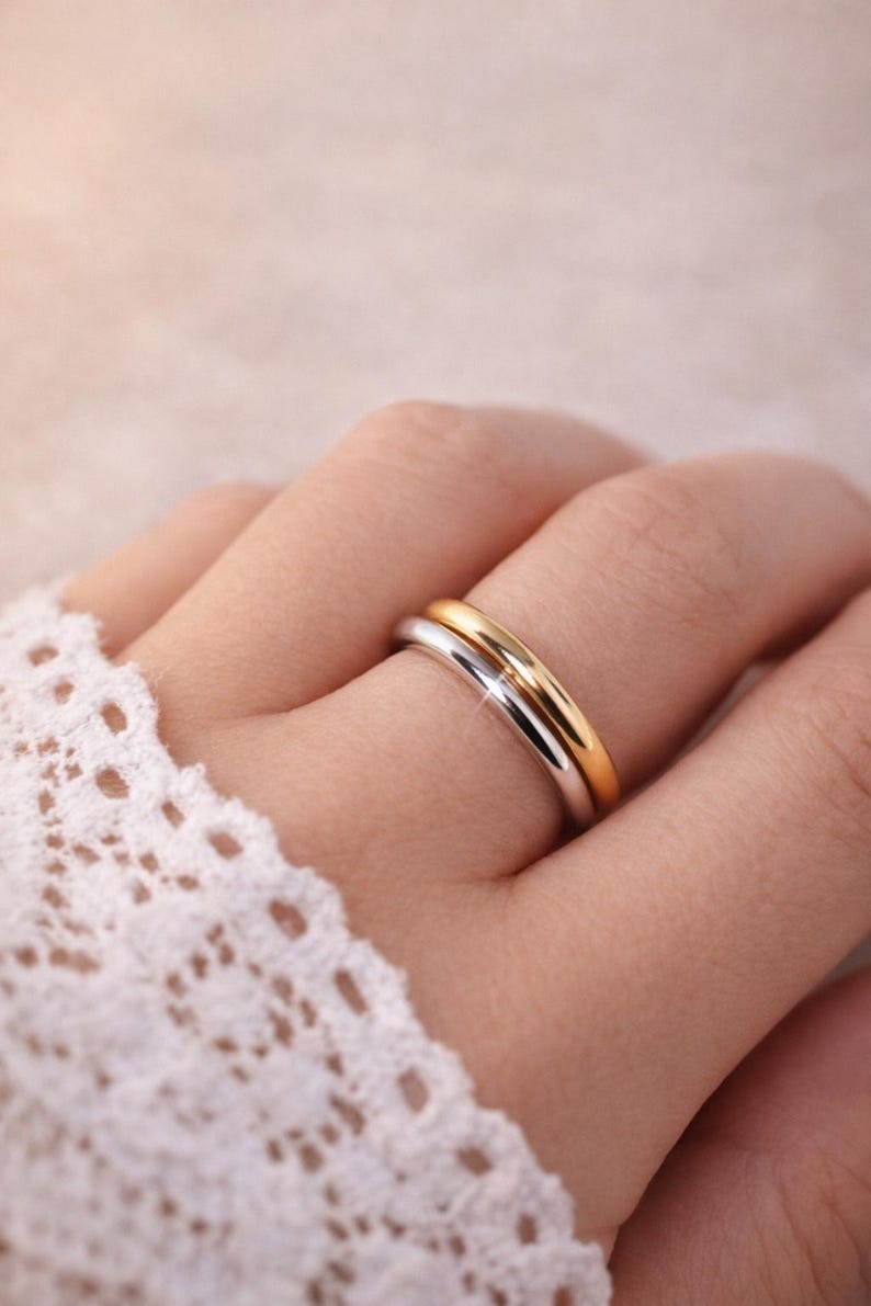 May include: A close-up of two rings, one gold and one silver, on a person's finger. The rings are smooth and shiny, with a simple band design. The background is soft and out of focus, with a hint of lace.