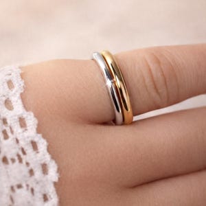 May include: Close-up of two rings, one silver and one gold, stacked on a finger. The rings are smooth and shiny, reflecting light. The background is blurred, focusing attention on the jewellery.