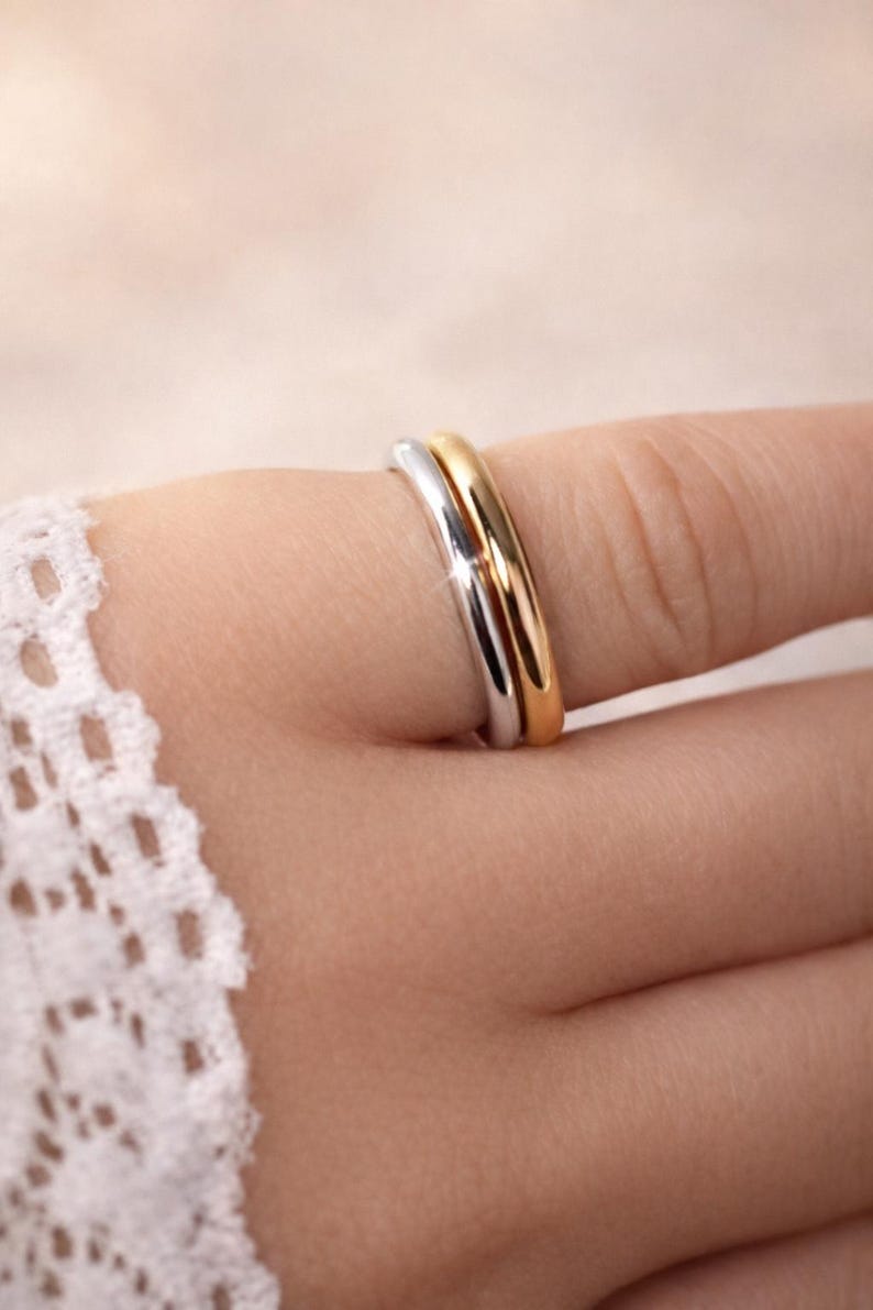 May include: Close-up of two rings, one silver and one gold, stacked on a finger. The rings are smooth and shiny, reflecting light. The background is blurred, focusing attention on the jewellery.