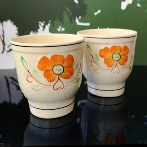 May include: Two white ceramic flower pots with orange and green floral designs. The pots are on a black surface.