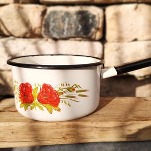 Op de afbeelding: Een witte emaille steelpan met een zwarte handgreep en rand. De steelpan heeft een bloemenontwerp met rode klaprozen en groen blad. De handgreep is zwart en wit. De steelpan staat op een houten oppervlak.