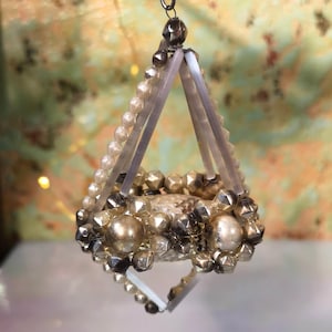 May include: A vintage-style ornament with a geometric design. The ornament features a combination of silver and pearl-like beads, with clear, rectangular supports. It hangs from a silver wire, showcasing a unique, handcrafted aesthetic.