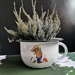 May include: A white enamelware pot with a black rim and handle, featuring a whimsical illustration of a hedgehog under an umbrella. The pot is filled with dried greenery. The background is black and green.