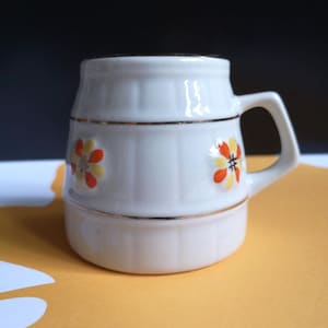 May include: A white ceramic mug with a gold rim and floral design. The flowers are yellow and orange with black centers.