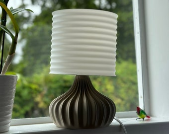 May include: A small, decorative table lamp with a white, horizontally ribbed lampshade. The base is a dark, ridged design. The lamp sits on a windowsill with a green, blurred background. A small, colorful bird figurine is visible.