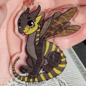 May include: A yellow and black dragon keychain with a clear acrylic background. The dragon has wings and is sitting on a small, detailed map.