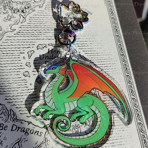 May include: Green and orange dragon charm with iridescent wings. The charm is hanging from a silver chain and is on top of a map with the text "Here Be Dragons".