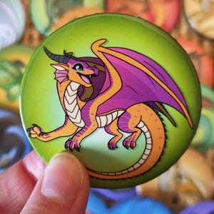 May include: A selection of colourful round badges with dragon illustrations. The central badge, held by a hand, features an orange dragon with purple wings on a green background. Other badges display various dragon designs.