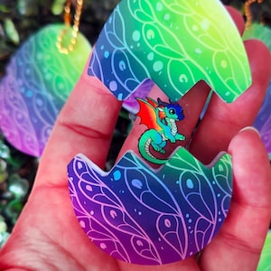 May include: A colourful dragon hatching from an egg-shaped pendant. The eggshell is decorated with a gradient of green, blue, and purple, with a delicate leaf pattern. The dragon is green, blue, and red. A gold chain is attached.