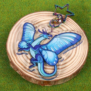 May include: A blue and purple dragon with butterfly wings, made of clear acrylic, is attached to a silver star-shaped keychain. The dragon is perched on a wooden circle.