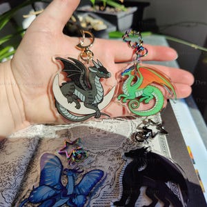 Op de afbeelding: Drie draken sleutelhangers met iriserende glitter. Een zwarte draak met een witte halve maan, een groene draak met oranje vleugels en een zwarte draak met een ster.