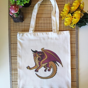 Puede incluir: Bolso de mano de lona blanca con un dise&ntilde;o de drag&oacute;n de dibujos animados. El drag&oacute;n es de color dorado, morado y marr&oacute;n. El bolso tiene dos asas blancas. El drag&oacute;n tiene grandes alas y sonr&iacute;e.
