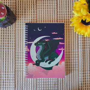 Peut inclure: Cahier à spirales avec un dragon vert sur un croissant de lune, sur fond de ciel nocturne nuageux. La couverture présente une palette de couleurs violet, rose et bleu. Le cahier est posé sur un tapis en bambou.