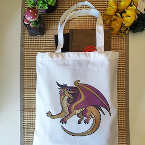 Puede incluir: Un bolso tote blanco con una ilustraci&oacute;n de un drag&oacute;n de dibujos animados en colores oro, morado y marr&oacute;n. El drag&oacute;n tiene grandes alas y est&aacute; en una pose din&aacute;mica. El bolso tiene asas blancas y contiene libros.