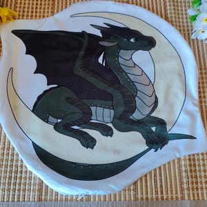 Könnte beinhalten: Ein weißes Kissen mit einer Drachenillustration. Der Drache ist grün und schwarz, mit einem hellgrauen Bauch, und sitzt auf einem Halbmond. Das Kissen liegt auf einer gewebten Matte mit gelben Blumen und einem kleinen dekorativen Pilz.