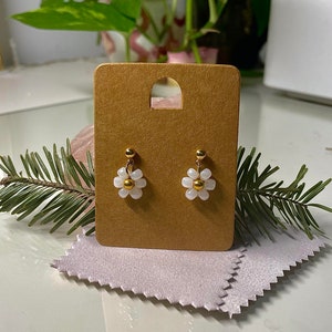 May include: A pair of gold and white flower stud earrings. The earrings are displayed on a brown card with a cutout in the centre.