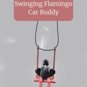 May include: A black bird-like figurine with pink legs and a white beak is seated on a pink swing. The swing is suspended by a black cord. The text "Swinging Flamingo Car Buddy" is displayed above the figurine.