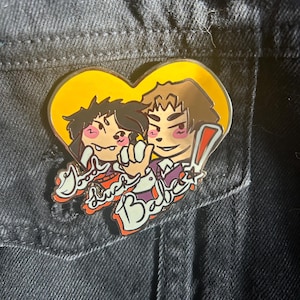May include: A gold heart-shaped enamel pin with two cartoon characters inside. The characters are a person with brown hair and a person with brown hair and a red bandana. The text "Good Luck Babe" is written in a stylized font below the characters.