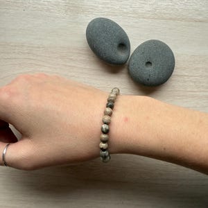 May include: A beaded bracelet with neutral-toned, round beads. The bracelet is worn on a wrist, with two gray stones in the background. The bracelet's beads have a mix of gray, brown, and tan colors.