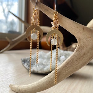 May include: Gold-tone earrings with a celestial design. Each earring has a crescent moon pendant, a small chain, and a round, white stone. The earrings also include a small, bone-like charm and a long chain that hangs below the moon.