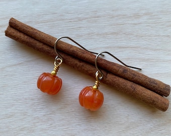 May include: A pair of orange pumpkin earrings with gold wire accents and silver hooks. The earrings are small and detailed, perfect for fall. The earrings are displayed on a light wood surface with two cinnamon sticks.