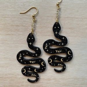 May include: A pair of black snake earrings with gold glitter, moons, and stars. The earrings have a gold hook and a crystal bead dangle.