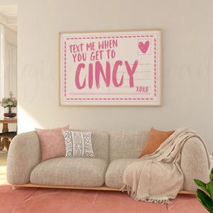 Puede incluir: Un estampado enmarcado con el texto "TEXT ME WHEN YOU GET TO CINCY" en rosa, con un corazón y "xoxo" en la parte inferior. El estampado cuelga sobre un sofá beige con cojines rosas y estampados, una manta melocotón y una alfombra rosa.