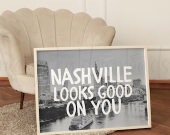 以下が含まれることがあります： 白黒のフレーム入りプリントで、白いフォントで「Nashville Looks Good On You」と書かれています。画像は川とボートのある都市景観を描いています。フレームは明るい木の色です。