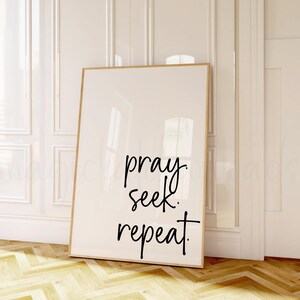 May include: A framed print with the words "pray. seek. repeat." in black script. The print has a light beige background and a thin, natural wood frame. The artwork is leaning against a white wall and door.