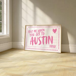 May include: A framed print with the text "TEXT ME WHEN YOU GET TO AUSTIN XOXO" in pink. The print has a postcard design with a heart and lines for writing. The frame is a light wood color.