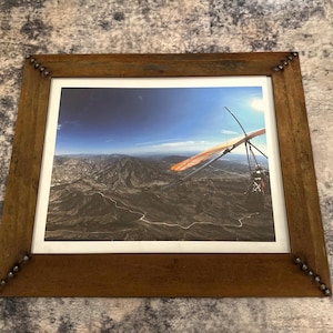 May include: A framed photograph of a hang glider soaring over a mountainous landscape. The frame is made of dark brown wood with metal corner accents. The image captures a wide view of the terrain under a blue sky.