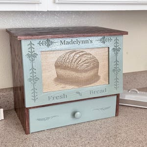 May include: A wooden bread box with a dark brown top and sides, featuring a light blue front panel. The panel displays the text Madelynn's Fresh Bread, a central illustration of a loaf of bread, and decorative floral carvings. A drawer is below.